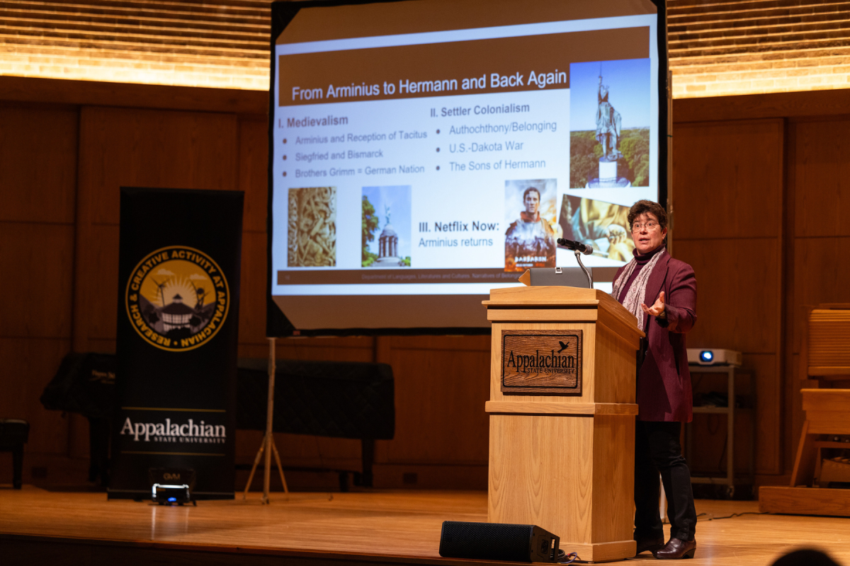 A speaker presents at a wooden podium in a university auditorium. Behind her, a projected slide titled “From Arminius to Hermann and Back Again” lists topics on medievalism, settler colonialism, and Netflix’s portrayal of Arminius, with related images. A banner beside the screen reads “Research & Creative Activity at Appalachian” and “Appalachian State University.”