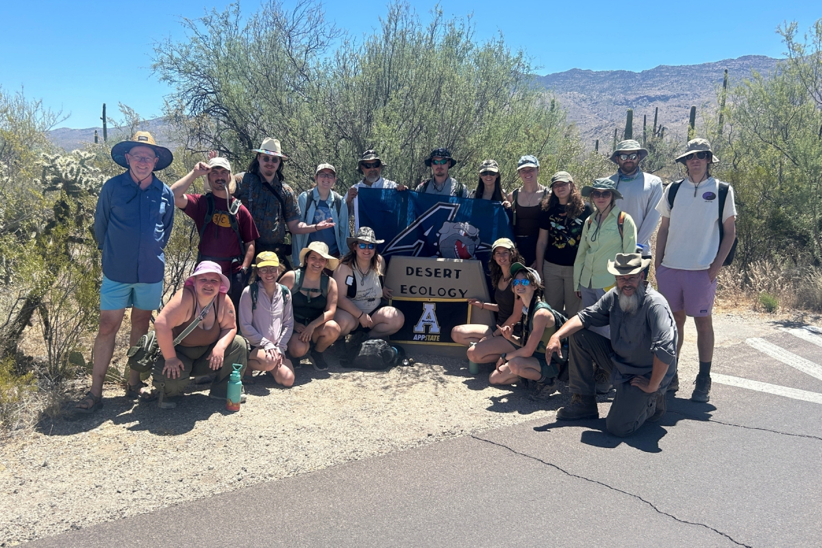 A group of people standing in a desert setting beside a sign reading "Desert Ecology." The group is holding flags featuring the Appalachian State and UNC Asheville logos.