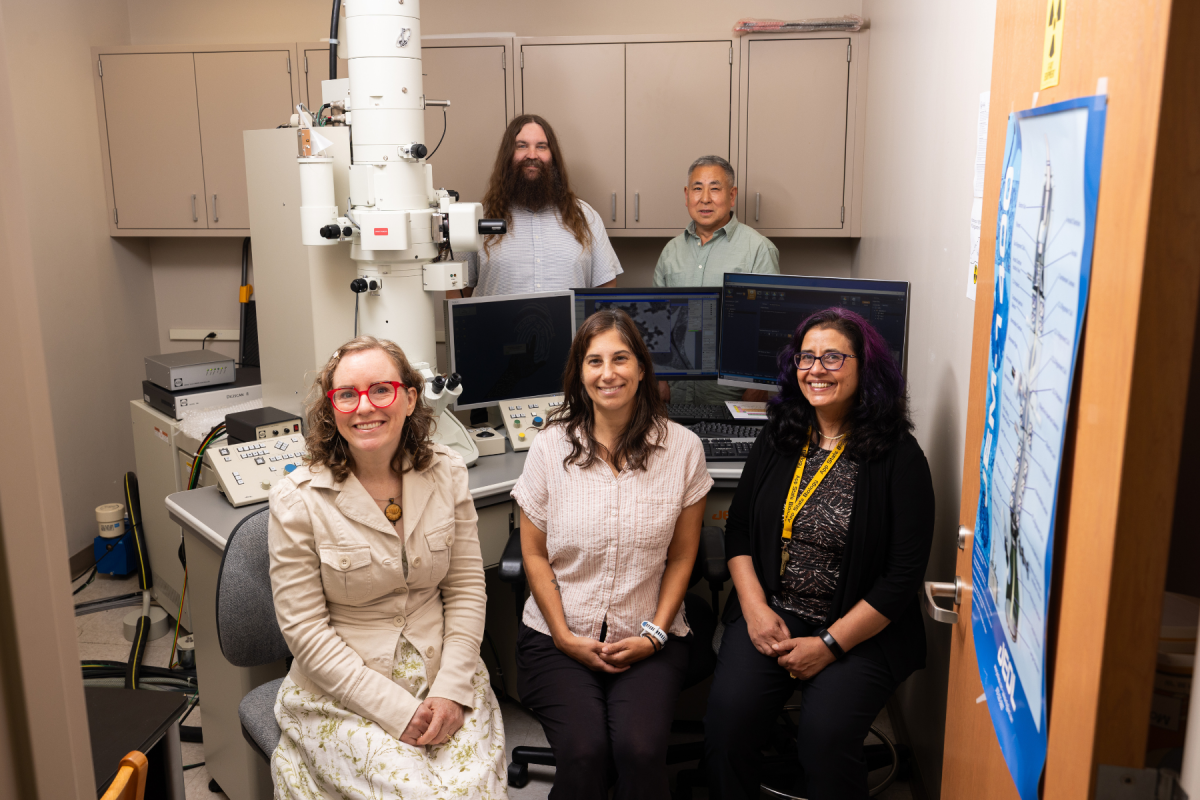 Five researchers pose in a laboratory beside a large microscope and computer monitors, smiling warmly in a collaborative, welcoming setting.
