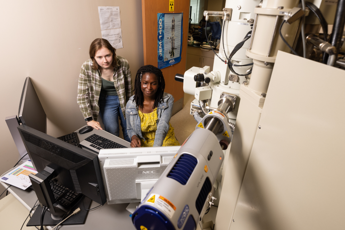 Two women examine computer monitors beside a large electron microscope in a lab, focused and collaborative during scientific research.