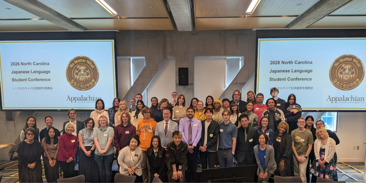 A large group of conference attendees poses together in front of two screens displaying the 2026 North Carolina Japanese Language Student Conference logo at App State&rsquo;s Plemmons Student Union.