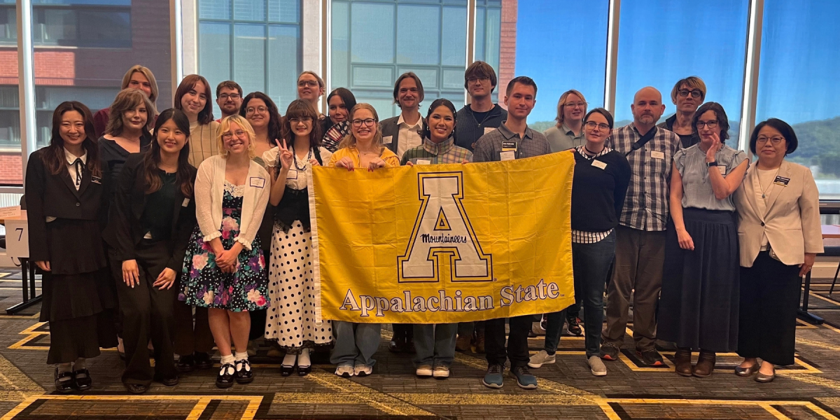 A group of App State conference participants poses indoors behind a large gold Appalachian State flag during the 2026 North Carolina Japanese Language Student Conference.