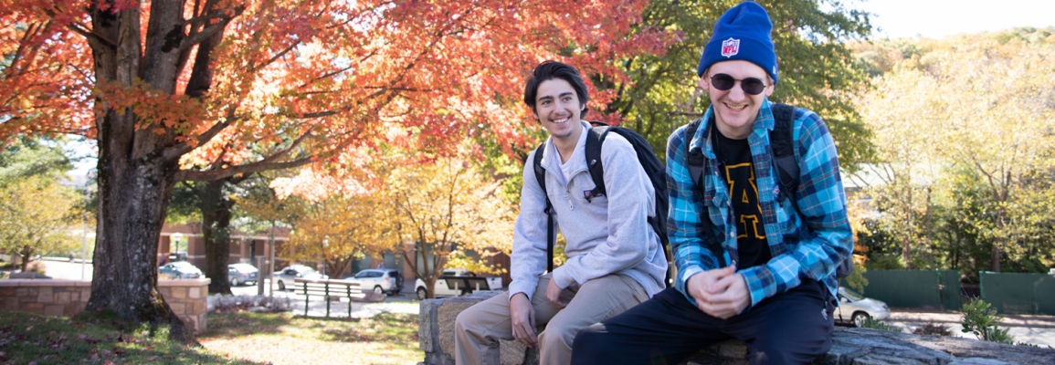 two male students sit on a rock wall, on campus with fall colors in the trees