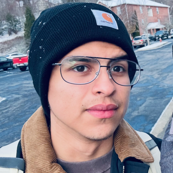 Close-up selfie of a person wearing glasses, a black beanie, and tan jacket outdoors near parked cars on a winter day.