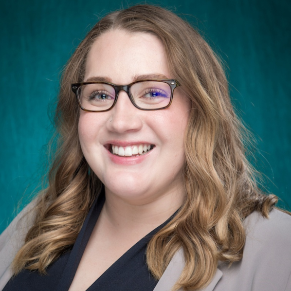 A headshot of College of Arts and Sciences Advising and Support Services Hub (CASSH) Academic Advisor and Student Records Coordinator Kaela Bray ’20 ’24. Bray is wearing glasses and a black shirt with a tan blazer. The background is bright blue.
