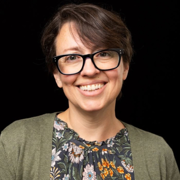 Smiling woman with short brown hair and black glasses, wearing a floral blouse and olive cardigan against a plain black background.