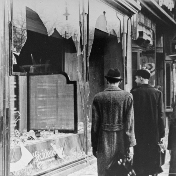 Black-and-white wartime street scene: two men walk past a bomb-damaged shop with shattered windows.