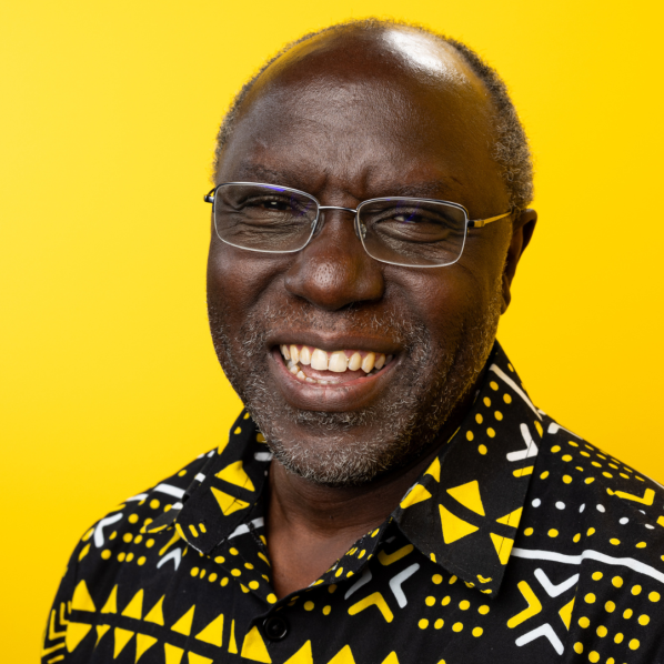 Smiling man with glasses and a beard, wearing a black shirt patterned with yellow geometric shapes against a bright yellow background.