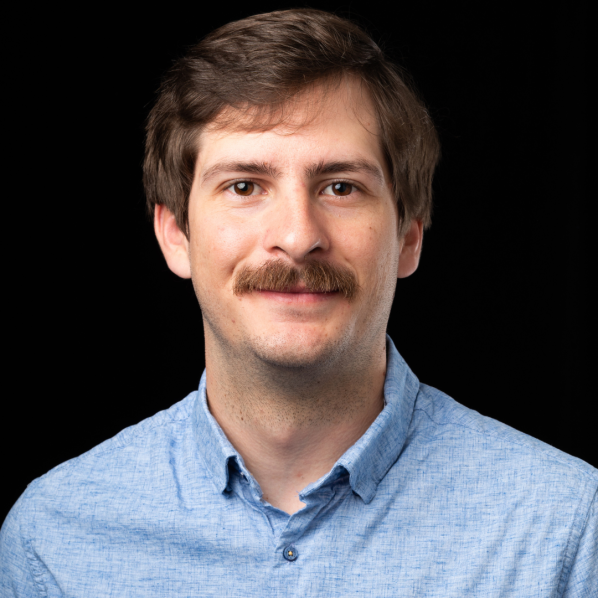A person with short, dark hair smiles while wearing a collared shirt, set against a black background.