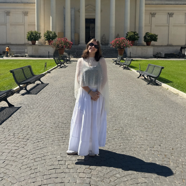 Woman in sunglasses and a flowing white outfit stands on a cobblestone path before a grand building, with benches, flowers, and bright sun.