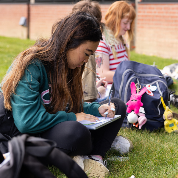 A group of people sitting on grass near a brick building. One person in focus is writing in a notebook, surrounded by a backpack with keychains. Others in the background are also engaged in various activities.