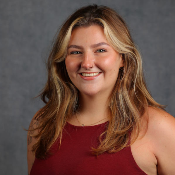 Smiling woman with long wavy blonde-brown hair, nose ring, and burgundy sleeveless top, posed against a gray studio background
