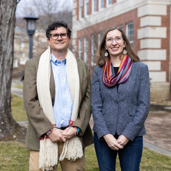 Two people stand outdoors in front of a brick academic building. The man on the left wears glasses, a brown blazer, light blue shirt, tan pants, and a cream-colored scarf with fringe. The woman on the right wears glasses, a gray blazer, dark jeans, and a multicolored scarf in shades of red, purple, and orange. Both are smiling, and the background shows a tree, grass, and a walkway.