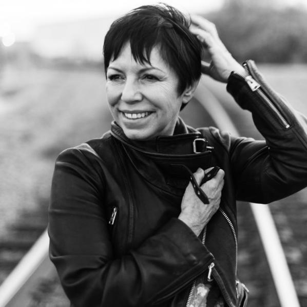 Black-and-white portrait of a smiling woman in a leather motorcycle jacket, touching her short hair outdoors.