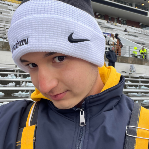 Close-up portrait of a person in a white beanie and winter jacket, standing in snowy stadium bleachers.