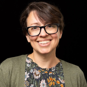 Smiling woman with short brown hair and black glasses, wearing a floral blouse and olive cardigan against a plain black background.