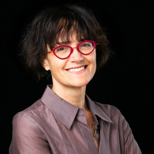 A person with short, dark hair and red glasses smiles while wearing a collared shirt, set against a black background.