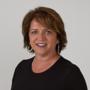 Professional headshot of a smiling woman with short brown hair, hoop earrings, and a black top against a neutral gray background.