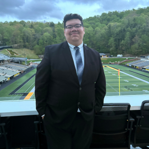 Smiling person in a black suit and tie stands in a stadium suite overlooking a football field, empty bleachers, and lush green hills.