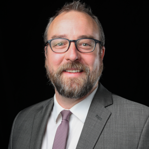 A person wearing a grey suit, white shirt, purple tie, and glasses smiles against a black background.