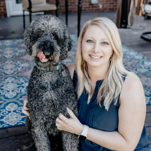 A woman sitting with a large, curly-haired dog on a patterned rug outdoors.