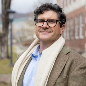Man with glasses and curly hair wearing a tan blazer, blue shirt, and cream scarf standing outside near a brick building.
