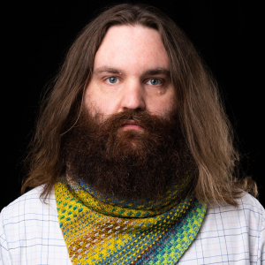 Studio portrait of a long-haired man with a thick beard, wearing a bright yellow, blue, and green knitted scarf over a checked shirt, against a black background.