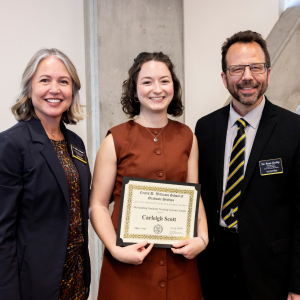 Three individuals smile for a photo. The middle individual is holding a certificate.