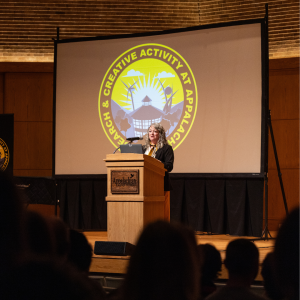 A woman stands at a wooden podium on stage in a university auditorium, speaking to an audience. Behind her, a large screen displays a circular yellow logo reading “Research & Creative Activity at Appalachian,” featuring stylized mountains, wind turbines, and a building under sunlight. The podium bears the Appalachian State University logo.