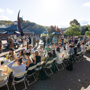 Outdoor community event with diverse guests dining at long tables, surrounded by art sculptures, greenery, and warm sunlight.