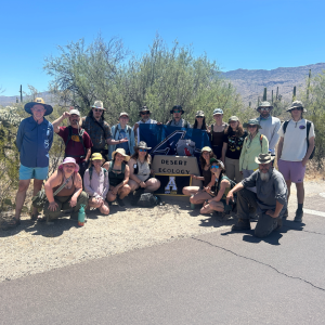 A group of people standing in a desert setting beside a sign reading "Desert Ecology." The group is holding flags featuring the Appalachian State and UNC Asheville logos.