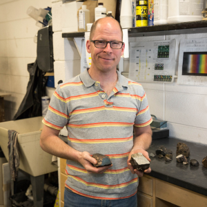 Smiling man in a striped polo holds two rock samples in a geology lab, with mineral specimens, charts, and equipment behind him.