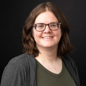 Studio portrait of a smiling woman with shoulder-length brown hair and patterned glasses, wearing a charcoal cardigan and olive top against a black background.